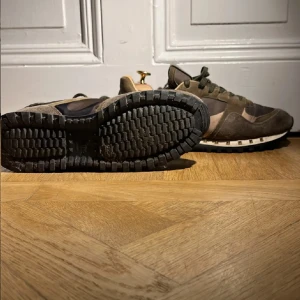 Valentino Rockrunner sneakers camouflage - Snygga Valentino Rockrunner sneakers i camouflage-mönster med olivgröna och bruna toner. Skorna har detaljer i mocka och textil, samt ikoniska nitar längs sulan. Grov yttersula och snörning framtill ger en cool och sportig vibe. Perfekta för dig som vill sticka ut. Ingen box 
