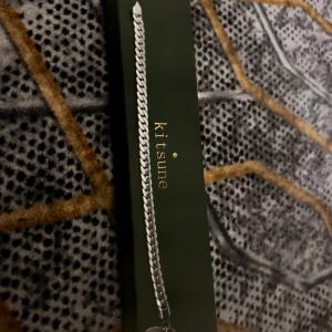 Silverfärgat armband från Kitsune - Stilrent armband i silverfärgad rostfri stål från Kitsune. Kedjan har en klassisk, platt länkdesign som ger en clean och modern look. Perfekt accessoar för att lyfta din outfit med en minimalistisk touch.