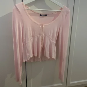 Ljusrosa volangblus från Gina Tricot - Supersöt ljusrosa blus från Gina Tricot med volangkant och liten rosett framtill. Blusen har lång ärm, croppad passform och spetsdetaljer vid halsringningen. Perfekt för dig som gillar romantisk och feminin stil.