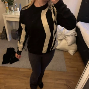 Svart och beige stickad tröja - Säljer en svart stickad tröja med breda beigea zigzag-ränder framtill och på ena ärmen. Tröjan har rund halsringning och långa ärmar. Perfekt för dig som gillar grafiska mönster och vill ha en snygg statement-piece i garderoben.