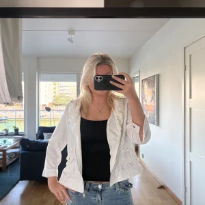 Vit jacka med detaljer  - Snygg vit jacka med dragkedja framtill och två fickor med lock och knapp. Jackan är kortare i modellen och har trekvartsärmar med knappdetalj vid ärmslutet. Står storlek 42 i men passar mer som S/M