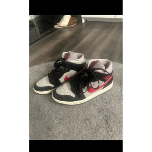 Nike Air Jordan 1 svart/vit/röd - Säljer ett par Nike Air Jordan 1 sneakers i svart, vitt och rött. Klassisk high-top modell med svart snörning, röd swoosh och detaljer, samt perforerad tå. Skorna har en ikonisk design och passar perfekt till streetwear.