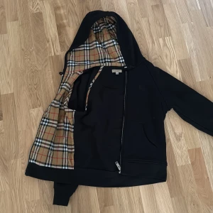 Svart hoodie Burberry - Svart hoodie från Burberry med rutig beige insida och huva, dragkedja framtill, två fickor och ribbade muddar. Två fläckar som inte syns om man inte vet om dem, se sista bilderna annars som ny, använd 2-3 gånger. 