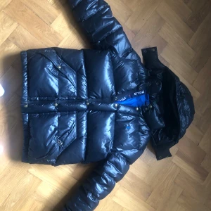 Svart pufferjacka från Polo Ralph Lauren - Snygg svart pufferjacka från Polo Ralph Lauren med glansig finish och huva. Jackan har dragkedja och knappar framtill, samt flera fickor med knappdetaljer. Perfekt för kalla dagar och har en cool, sportig look.