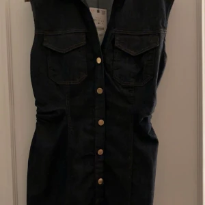 Svart jeansklänning från Zara M - Snygg svart jeansklänning från Zara i storlek M. Klänningen är ärmlös med markerad midja, två bröstfickor och knappar framtill i guldton. Perfekt för dig som gillar en cool och stilren look. Aldrig använt bara testat, passar till en tjej med större byst