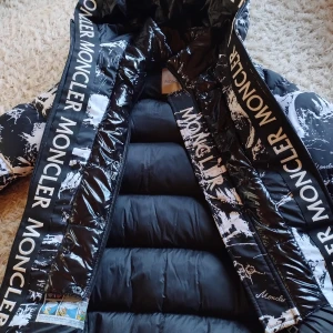 Svartvit Moncler dunjacka med tryck - Snygg svartvit dunjacka från Moncler med coolt abstrakt mönster och stora loggor på insidan av huvan och dragkedjan. Jackan har flera dragkedjor framtill och är vadderad för extra värme. Perfekt för dig som vill sticka ut i vinterkylan. Otroligt varm. Endast Använd fåtal ggr. Xs-s