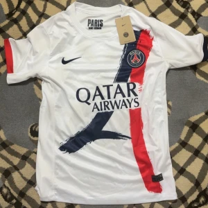 PSG  fotbollströja - Säljer en snygg Paris Saint-Germain fotbollströja från Nike i vit polyester med tryck i marinblått och rött. Tröjan har korta ärmar, PSG-logga på bröstet och Qatar Airways som sponsor. Perfekt för dig som älskar fotboll och PSG.