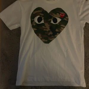 Vit Comme des Garçons Play t-shirt - Säljer en vit t-shirt från Comme des Garçons Play med stort hjärta i camouflage och ögon på bröstet. Liten röd hjärtlogga på hjärtat. Klassisk passform och rund hals. Perfekt statement-plagg för dig som gillar streetwear.