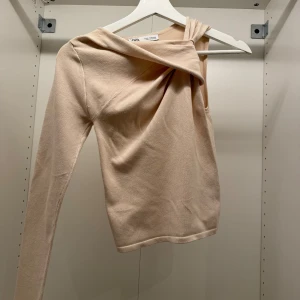 Beige oneshoulder topp från Zara - Säljer en beige oneshoulder topp från Zara med lång ärm och snygg drapering över axeln. Toppen har en stilren och modern look med asymmetrisk halsringning. Mjuk och skön kvalitet som passar perfekt till din garderob.