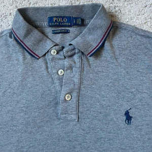 Grå pikétröja från Polo Ralph Lauren - Snygg grå pikétröja från Polo Ralph Lauren med klassisk krage och knäppning framtill. Tröjan har marinblå och vinröda detaljer på krage och ärmslut samt den ikoniska broderade loggan på bröstet. Modellen är slim fit och i mjuk bomull. Storlek s men sitter xs (slim)