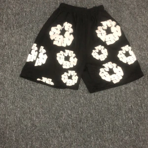 Svarta shorts från Denim Tears med tryck - Svarta shorts från Denim Tears med elastisk midja och stora vita grafiska tryck i form av kransar över hela plagget. Sköna och loose fit, perfekta för en chill och trendig look. Materialet känns som bomull och shortsen har en avslappnad vibe.