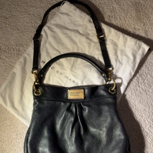  Äkta Svart axelväska Marc by Marc Jacobs - Snygg svart axelväska från Marc by Marc Jacobs i mjukt skinn med guldfärgade metalldetaljer och klassisk logoplatta framtill. Väskan har både handtag och justerbar axelrem, perfekt för att bära på olika sätt. Kommer med original dustbag.