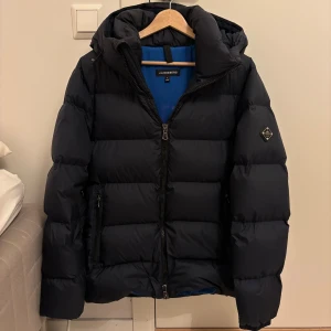 Svart dunjacka från J.Lindeberg - Snygg marin blå dunjacka från J.Lindeberg med huva och dragkedja framtill. Jackan har en puffig design, diskret logga på ärmen och två fickor med dragkedja. Perfekt för kalla dagar när du vill hålla stilen och värmen. Jackan är använd ett fåtal gånger förra vintern. 