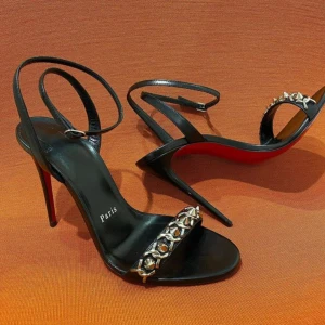 Svarta sandaler med klack och nitar - Snygga svarta sandaler från Christian Louboutin med hög klack och röd sula. Skorna har smala remmar runt vristen och en dekorativ rem framtill med silvriga nitar. Tillverkade i skinn för en lyxig känsla och edgy look.