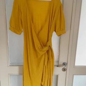 Gul omlottklänning från ASOS - Säljer en lång gul omlottklänning från ASOS med puffiga korta ärmar och knyt i midjan. Klänningen har djup v-ringning och är tillverkad i ett mjukt bomullsmaterial. Perfekt för dig som gillar färg och vill sticka ut.