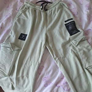 Ljusgröna cargopants med snörning - Snygga ljusgröna cargopants med elastisk midja och svart snörning. Byxorna har stora fickor på sidorna och coola svarta patchar framtill. Tillverkade i mjuk bomull, perfekta för en avslappnad streetstil. Det finns en gul fläck på övre delen av låret på vänstee sida. 