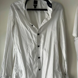 Vit slim fit skjorta från H&M XS - Snygg vit skjorta från H&M i slim fit-modell, tillverkad i premium bomull. Skjortan har svarta knappar och lång ärm. Insidan av kragen har ett marinblått tyg med små vita mönster för en extra detalj. Perfekt för dig som gillar stilrena plagg med en twist.