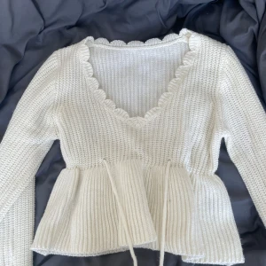 Vit stickad blus med volangkant - Supersöt vit stickad blus med v-ringning och volangkant längs halsen. Modellen har långa ärmar, knytband i midjan och en peplum-liknande nederdel. Perfekt för lager-på-lager och ger en gullig touch till din outfit.