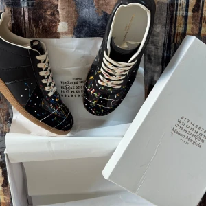 Maison Margiela sneakers med färgstänk - Maison Margiela sneakers i svart skinn med coola färgstänk i blått, rött, gult och vitt. Skorna har vita snören, beige innersula och klassisk gummisula. Unik design som sticker ut och passar perfekt för dig som gillar statement pieces.