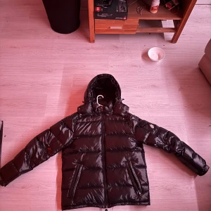 Svart Moncler pufferjacka med huva - Säljer en svart, glansig pufferjacka från Moncler med huva och dragkedja. Jackan har två snedställda fickor framtill och en ärmficka med Moncler-logga. Insidan har det klassiska Moncler-serietrycket. Perfekt för kalla dagar och riktigt snygg streetstil.