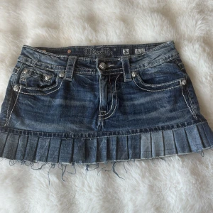 Miss Me jeanskjol med broderi och nitar - Säljer en blå jeanskjol från Miss Me med plisserad nederkant och råa fransar. Kjolen har snygga broderade bakfickor med silverdetaljer och nitar samt ett stort lädermärke med metallkors bak. Klassisk femficksmodell och låg midja.