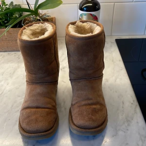 Bruna UGG boots med fårskinn - bruna UGG boots med mjukt fårskinnsfoder och platt sula. Klassisk rund tå och robust mocka på utsidan. Perfekta för kalla dagar och har en enkel, tidlös design med UGG-logga bak på hälen.
