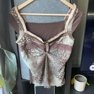 Brun och beige linnetopp - Säljer en brun och beige linnetopp med djurmönster framtill och enfärgad baksida. Toppen har spetsdetaljer vid axelbanden och en liten rosett framtill. Materialet känns mjukt och lätt, perfekt för varma dagar. Snyggt fall och breda axelband. Står storlek XXL och 100% polyester