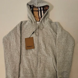 Burberry hoodie / zip  - Snygg grå hoodie från Burberry med klassiskt rutigt foder i huvan och dragkedja framtill.    Helt ny, säljer för att den passade inte mig. Storlek : S. Hör av er för mer info, priset kan diskuteras vid snabb affär.