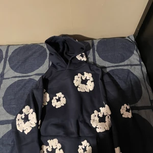 Mörkblå hoodie från Denim Tears - Säljer en mörkblå hoodie från Denim Tears med vita blommönster över hela tröjan. Hoodien har huva och en klassisk känguruficka framtill. Perfekt för dig som vill sticka ut med en unik och trendig streetwear-look.
