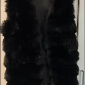 Svart fluffig pälsväst - Säljer en svart pälsväst med riktigt fluffig och mjuk känsla. Västen har en rak passform och är perfekt för att ge din outfit en extra touch. Den är helsvart och har en stilren look som funkar till många olika stilar.
