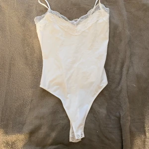 Vit spetskantad bodysuit från Bershka - Säljer en vit bodysuit från Bershka med tunna axelband och v-ringad spetskant upptill. Bodyn har tryckknappar i grenen och är tillverkad i mjuk bomull med stretch. Perfekt för att styla med jeans eller kjol för en clean look.