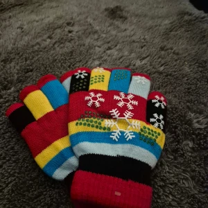 Färgglada stickade vantar med snöflingor - Säljer ett par riktigt färgglada stickade vantar med snöflingor på ovansidan. Vantarna har ränder i rött, gult, blått, svart och ljusblått samt små gröna prickar. Perfekta för vintern och julen när du vill sticka ut lite extra!