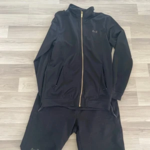 Svart träningsjacka från A–Z, stl L - Svart träningsjacka från A–Z med guldfärgad dragkedja och diskret logga på bröstet. Jackan har hög krage, två fickor med dragkedja och är tillverkad i 100% polyester. Perfekt för dig som gillar sportig och clean stil.