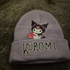 Lila Kuromi mössa - Säljer en söt lila stickad mössa med Kuromi-tryck framtill och texten 'Kuromi' på uppviket. Perfekt accessoar för dig som gillar kawaii-stil och vill sticka ut lite extra.