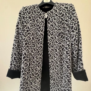 Vändbar svartvit ullkappa - Betsey Johnson - Snygg ullkappa som är svartvit på ena sidan och helt svart då man vänder insidan ut. Betsey Johnson. Jackan har rund hals med en stilren metallknäppning framtill. Perfekt för större halsdukar när det blir kallt. Längd från axel och ner ca 82 cm.