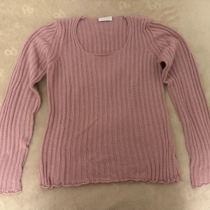 stickad tröja - Supersöt ljusrosa stickad tröja från Versi med ribbat mönster och långa ärmar. Tröjan har en rundad halsringning och fina volangdetaljer längst ner. Perfekt för lager-på-lager och passar till både jeans och kjol.