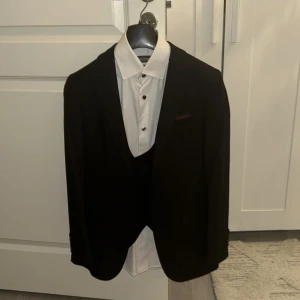 Svart kostym från Cengiz Inler - Säljer en stilren svart kostym från Cengiz Inler med matchande väst, vit skjorta och svart slips. Kavajen har klassiska slag och en vinröd näsduk i bröstfickan. Kostymen är i polyester och levereras i original fodral. Perfekt för dig som vill vara extra snygg på bal eller fest. Använd 1 gång.