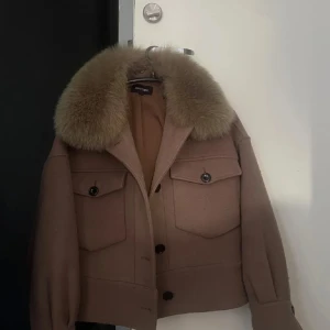 Meotine kappa - Säljer en beige jacka från Meotine med stor fluffig pälsig krage. Jackan har två stora bröstfickor med knappar, markerade axlar och knäppning framtill. 