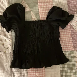 Svart blus med puffärmar från Seaside - Säljer en svart blus från Seaside med bred smockad överdel och stora puffärmar. Blusen har en fyrkantig halsringning och är tillverkad i ett mjukt, lätt material som ger en snygg siluett. Perfekt för dig som gillar trendiga detaljer och volym.