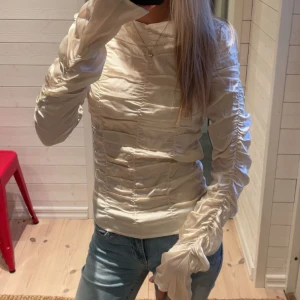 Champagnefärgad sidenblus med rynk - Säljer en champagnefärgad blus i siden med coola rynkade detaljer över hela toppen och ärmarna. Blusen har lång ärm och en elegant, glansig finish.💕