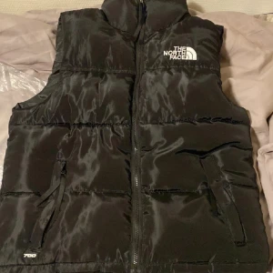 Svart dunväst från The North Face - Svart dunväst från The North Face med hög krage, dragkedja framtill och två fickor med dragkedja. Klassisk logga på bröstet och 700-brodyr vid nederkanten. Perfekt för lager-på-lager och streetwear.