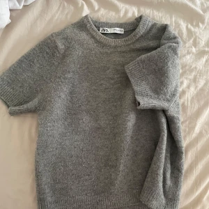 Grå stickad tröja från Zara - Mysig grå stickad tröja från Zara med korta ärmar och rund halsringning. Tröjan har en enkel och stilren design med ribbade kanter vid hals, ärmslut och nederkant. Perfekt att passa på att köpa den nu under hösten eller våren. Har blivit använd ganska mycket och har lite smink runt halsen. Är i storlek M men passar mig som är i XS/S