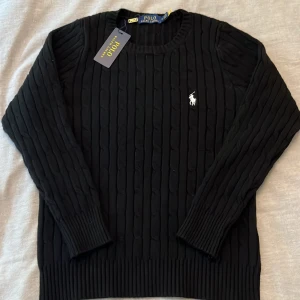 Svart kabelstickad tröja från Polo Ralph Lauren - Säljer en kabelstickad svart tröja från Polo Ralph Lauren med klassisk rund halsringning och broderad vit logga på bröstet. Nyskick, inga defekter. Perfekt inför kallare höstdagar 🍁Hör av er vid frågor 🙌