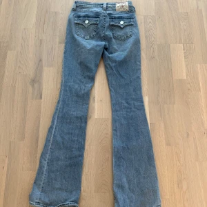 True Religion Becca Bootcut Jeans - Snygga ljusblå bootcut jeans från True Religion, modell Becca Mid Rise. Klassiska femficksdetaljer, markerade sömmar och ikoniska bakfickor med lock och silverfärgade knappar. Jeansen är i mjuk bomullsdenim och har en riktigt schysst passform.