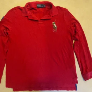 Röd långärmad pikétröja Polo Ralph Lauren - Röd långärmad pikétröja från Polo Ralph Lauren med klassisk krage och stor broderad polospelare på bröstet. Tröjan är i mjuk bomull och har en avslappnad passform, perfekt för en stilren och sportig look.