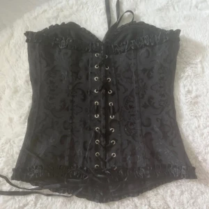 Svart korsett med snörning och mönster - Säljer en svart korsett med vackert blommigt mönster och glansig finish. Toppen har snörning framtill, volangkant upptill och nertill samt satinband som detalj. Perfekt nu till halloween. Min kamera är sönder där av lite dåliga bilder!