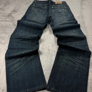 Mörkblå jeans Sarol 858 wide fit - Säljer ett par mörkblå jeans från Sarol 858 med breda ben och snygga slitningar. Jeansen har klassiska fem fickor, kontrastsömmar och broderad detalj på bakfickan. Perfekta för dig som gillar en loose och avslappnad stil.