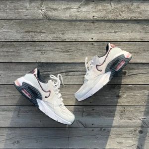 Nike Air Max vita sneakers - Nike Air Max sneakers i vitt med mörkblå och vinröda detaljer. Skorna har en chunky sula med synlig Air Max-dämpning, vit ovandel i mesh och skinn samt broderad Swoosh i vinrött. Klassisk snörning och svart innersula. Perfekta för dig som gillar streetstyle.