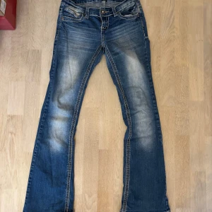 Blåa bootcut jeans från Anya Jeans - Snygga blåa bootcut jeans från Anya Jeans med slitningar och kontrastsömmar. Bakfickorna har broderade detaljer och paljetter i blått och silver. Jeansen har klassisk femficksdesign och markerad tvätt för en cool look. Storlek 36!🩷