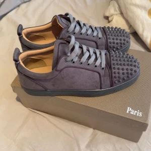 Christian Louboutin sneakers med nitar - Snygga mörkgrå sneakers från Christian Louboutin i mocka med ikoniska röda sulor och coola nitar på tån. Skorna har grå snörning och en platt sula. Perfekta för dig som vill sticka ut med en lyxig och edgy stil.